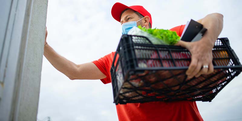 Homem de uniforme vermelho segurando uma caixa de produtos enquanto toca a campanhia de uma casa