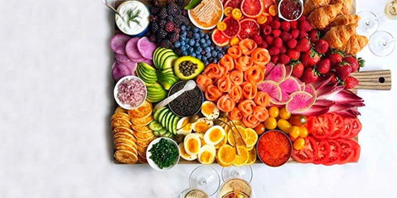 Tábua quadrada repleta de diferentes tipos de alimentos ao centro de uma mesa branca