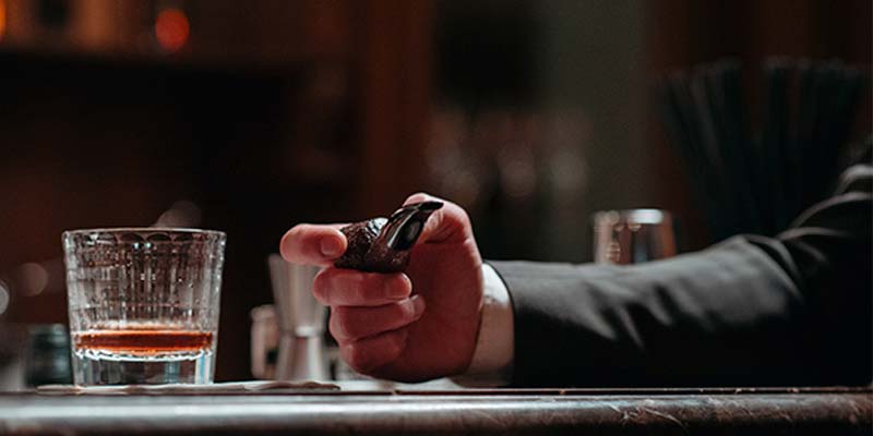 Homeme de terno segurando um cachimbo em uma mesa de bar com um copo de Whisky